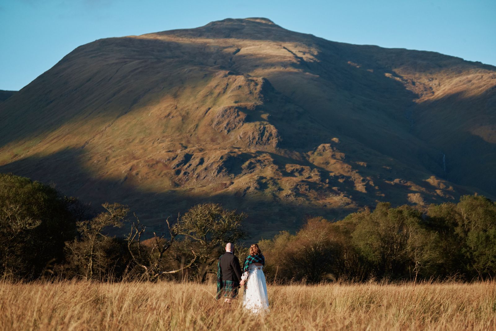 A couple in a golden meadow beneath a sunlit Highland mountain