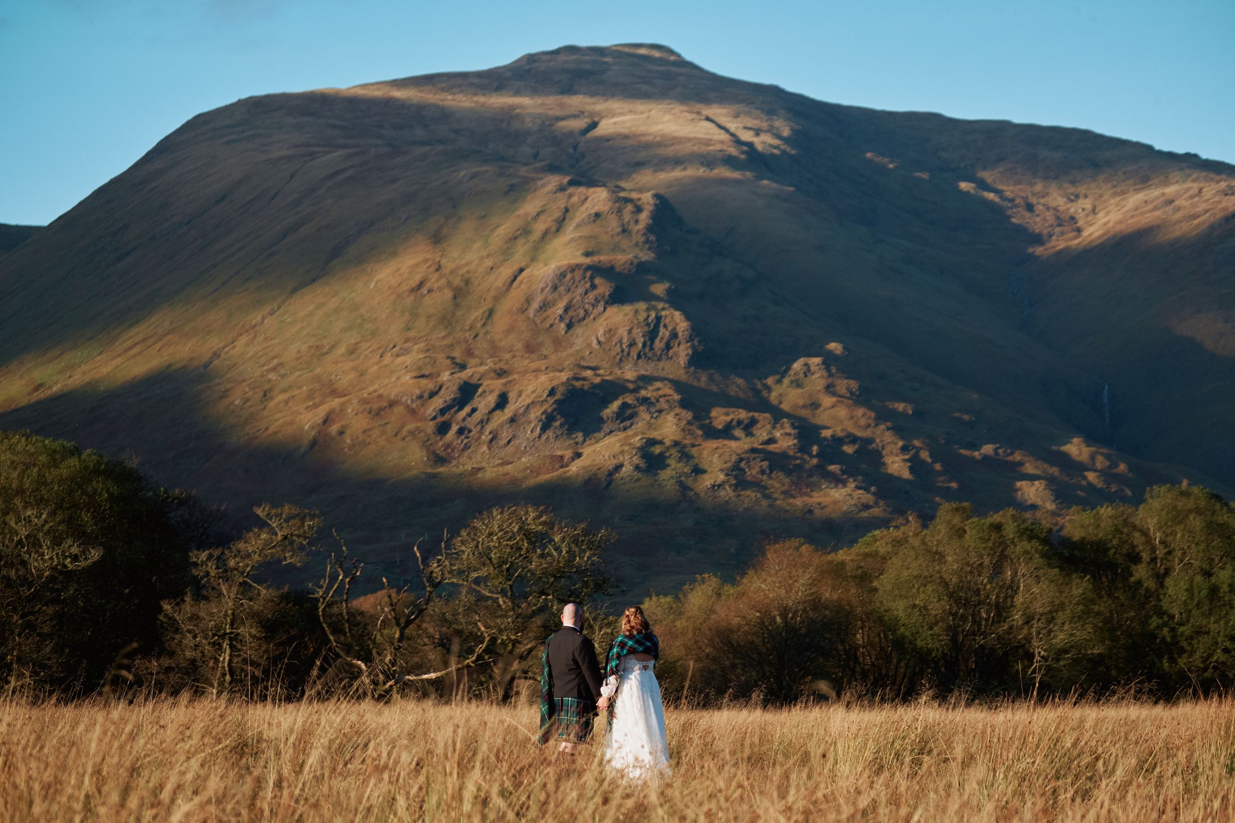 A couple in a golden meadow beneath a sunlit Highland mountain