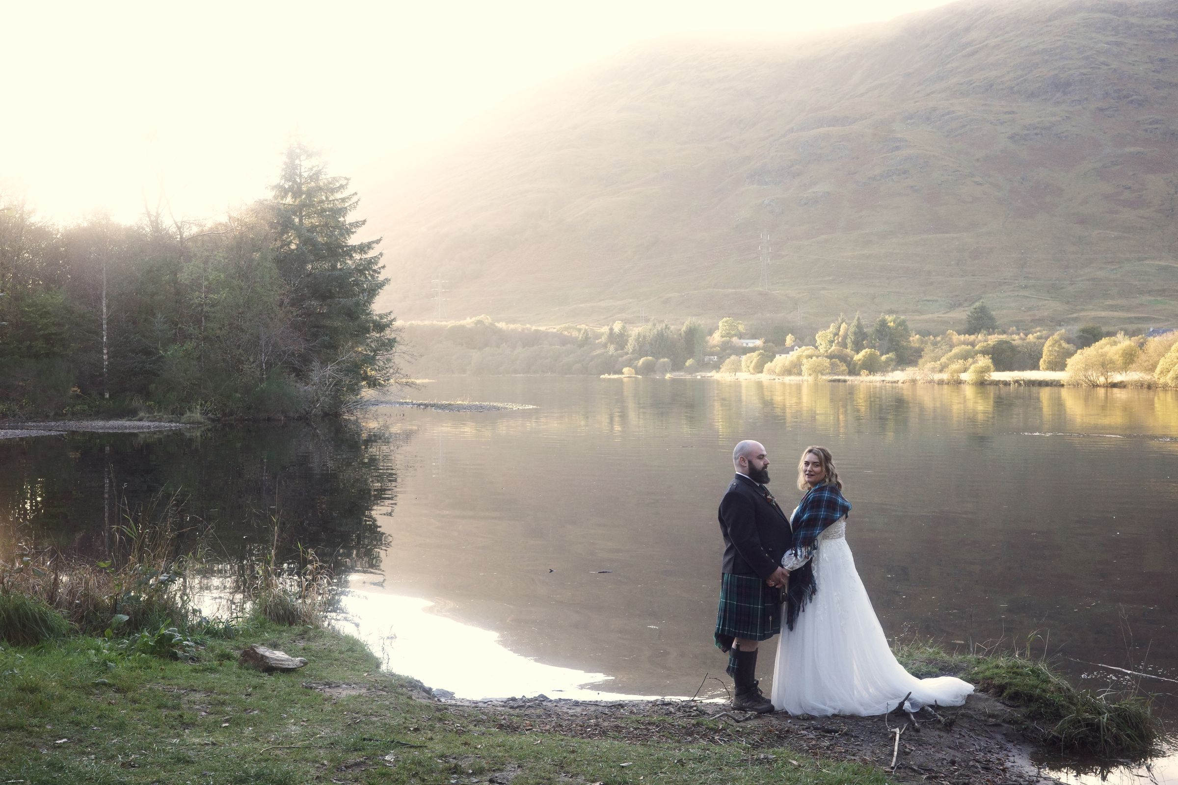 A couple standing at the edge of a misty loch in golden morning light